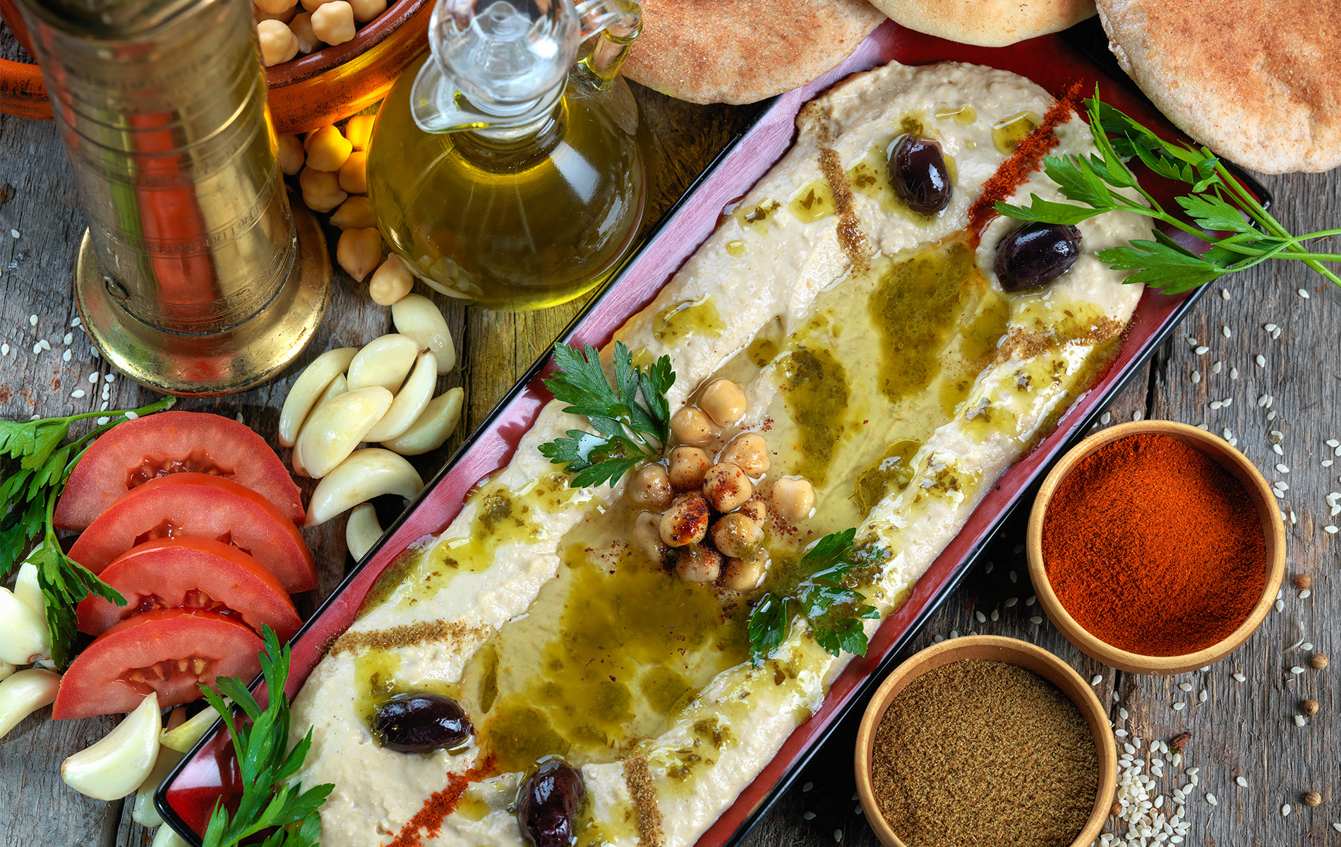 Large hummus platter on wooden table surrounded by pita bread and other fresh ingredients.