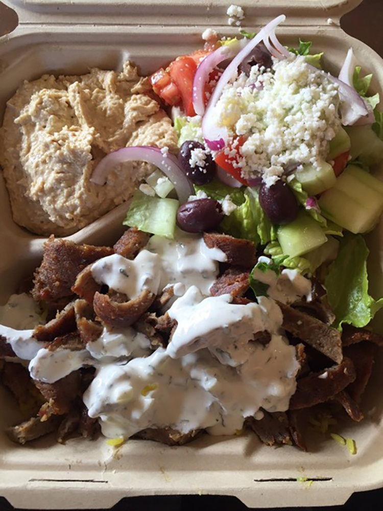 A to-go paper box with a serving of Hummus, Greek Salad, Gyro meat with a Tzatziki sauce.